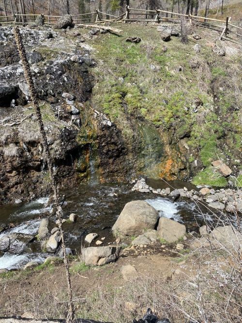 A small waterfall cascades over rocky terrain, surrounded by green grass and a flowing stream below.