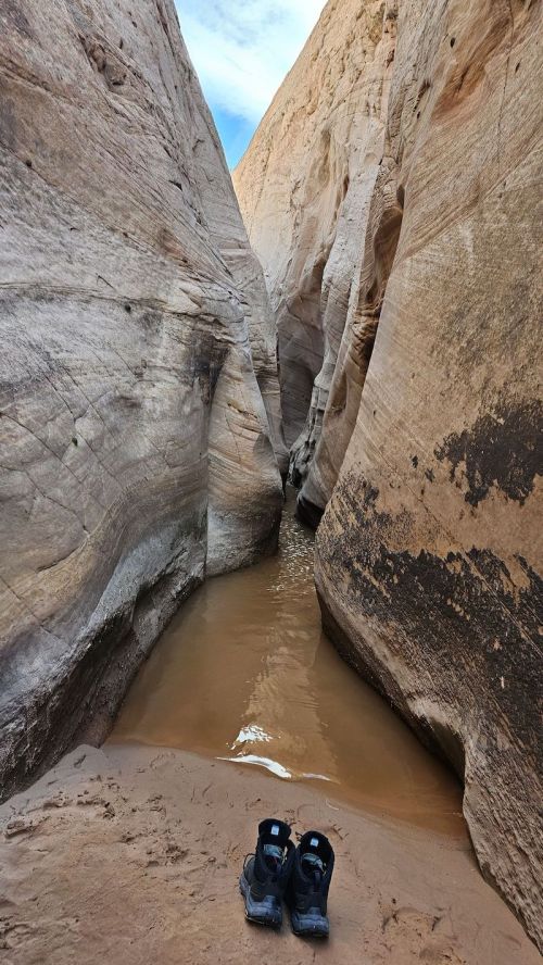 A narrow canyon with smooth rock walls and a small pool of water, featuring a pair of black shoes on the sandy ground.