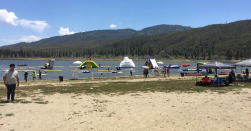 A sandy beach by a lake with inflatable water slides, people enjoying the sun, and mountains in the background.