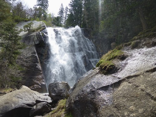 A cascading waterfall flows over large rocks, surrounded by lush greenery and tall trees in a serene natural setting.