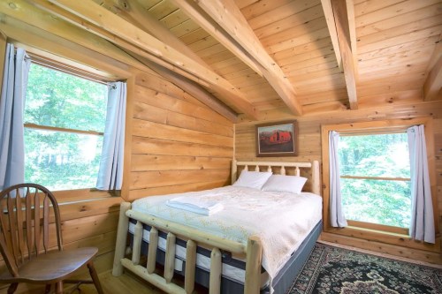 Cozy wooden bedroom with a log bed, two windows, a chair, and a rustic painting on the wall.
