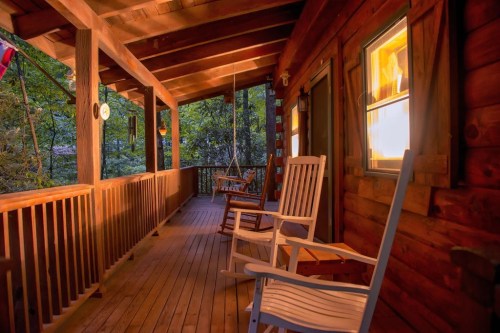 A cozy wooden porch with rocking chairs, illuminated by warm light, surrounded by trees in the evening.