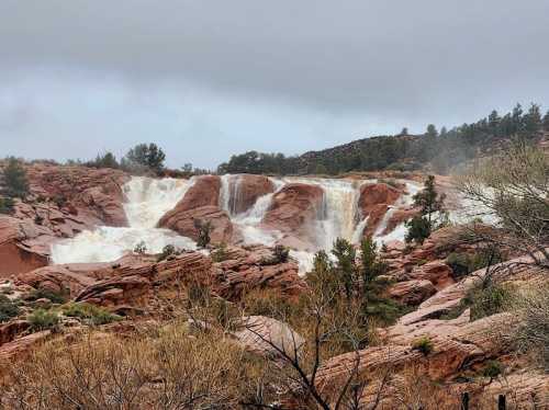 A waterfall cascades over red rock formations, surrounded by sparse vegetation and a cloudy sky.