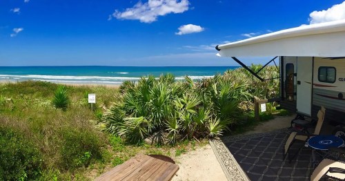 A scenic view of a beach with turquoise water, palm trees, and an RV under a blue sky with fluffy clouds.