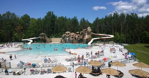 A large outdoor pool with water slides, surrounded by trees and people enjoying the sun on lounge chairs.