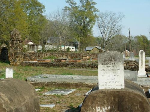 A cemetery with old gravestones, surrounded by trees and distant buildings under a clear sky.