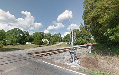 A railroad crossing with a signal, surrounded by trees and a rural landscape under a partly cloudy sky.