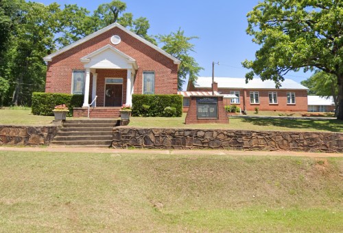 A brick building with white trim, surrounded by greenery, featuring steps and a sign in front on a sunny day.
