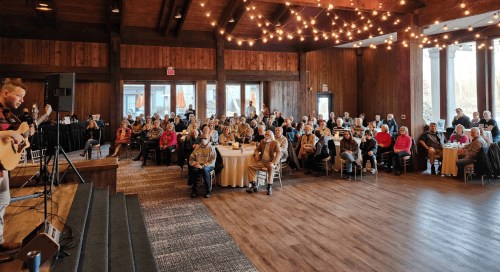 A musician performs on stage for a large audience seated at tables in a warmly lit venue with wooden decor.