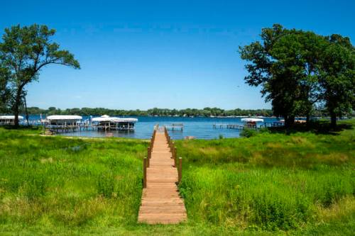 A wooden dock leads to a serene lake surrounded by green grass and trees under a clear blue sky.