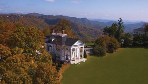 A grand white mansion surrounded by colorful autumn trees and rolling mountains under a clear blue sky.