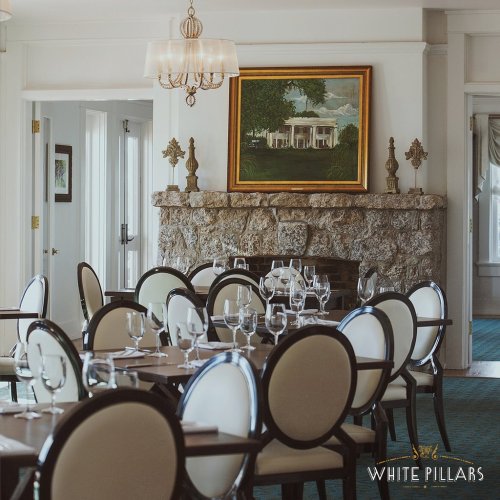 Elegant dining room with round tables, white chairs, and a large painting above a stone fireplace. Bright and inviting atmosphere.