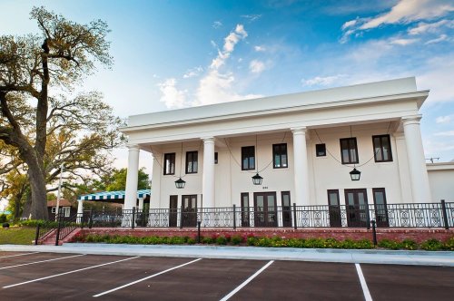 A modern building with columns, large windows, and a landscaped parking area under a blue sky.