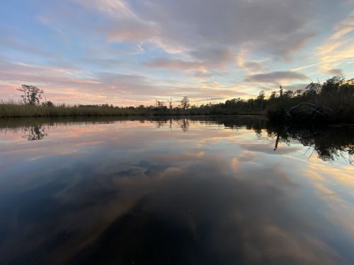A serene river scene at sunset, with colorful clouds reflected in the calm water and trees lining the shore.