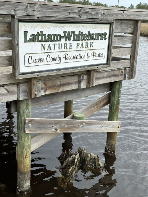 Sign for Latham-Whitehurst Nature Park on a wooden pier over water, with a tree stump visible below.