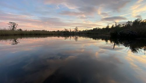 A tranquil water scene at sunset, with colorful clouds reflecting on the calm surface surrounded by trees.