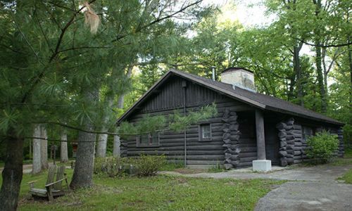 A rustic log cabin surrounded by trees, with a stone chimney and a path leading to the entrance.