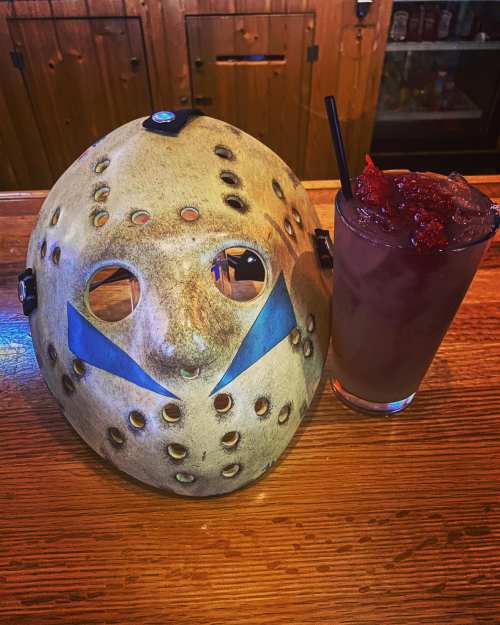 A hockey mask with blue markings next to a cocktail garnished with red fruit on a wooden bar.