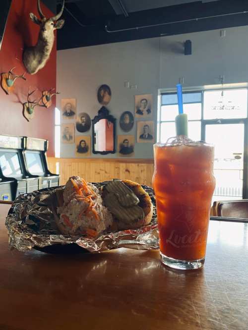 A sandwich with pickles and a drink on a bar table, with vintage photos and a deer head on the wall in the background.