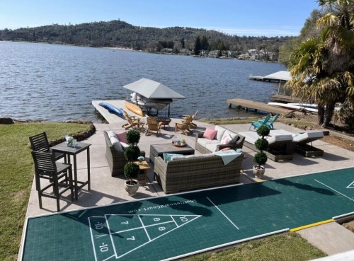 A lakeside patio with seating, a table, and a shuffleboard court, surrounded by greenery and water views.