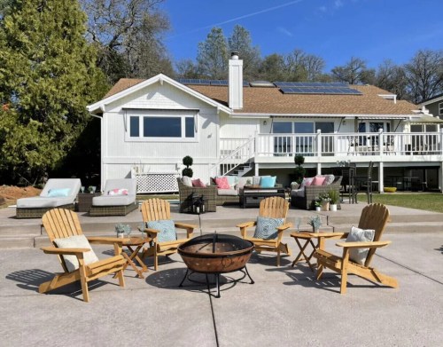 A cozy outdoor seating area with wooden chairs and a fire pit, set in front of a white house with a deck and solar panels.