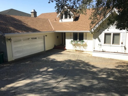 A white house with a brown roof and a garage, surrounded by trees and a paved driveway.