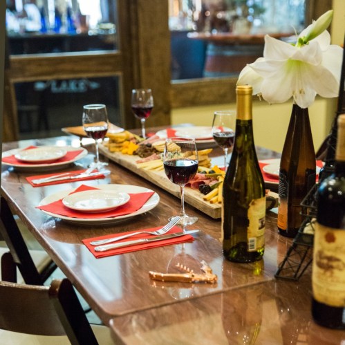 A dining table set with red napkins, wine glasses, and a platter of food, featuring bottles of wine and a flower centerpiece.