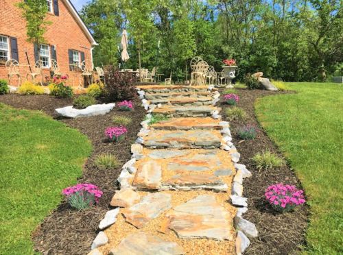 A stone pathway leads through a landscaped garden with flowers, grass, and outdoor seating near a brick house.