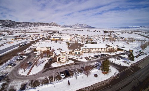 Aerial view of a snowy town with mountains in the background, featuring buildings, roads, and parked cars.