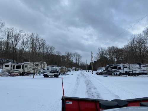 A snowy RV park with parked trailers and vehicles, surrounded by bare trees under a cloudy sky.