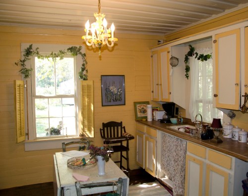 Cozy kitchen with yellow walls, a chandelier, a window, and a wooden table adorned with flowers and kitchenware.