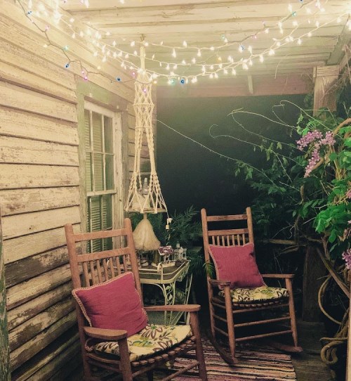Cozy porch with two rocking chairs, colorful cushions, string lights, and lush greenery in a rustic setting.