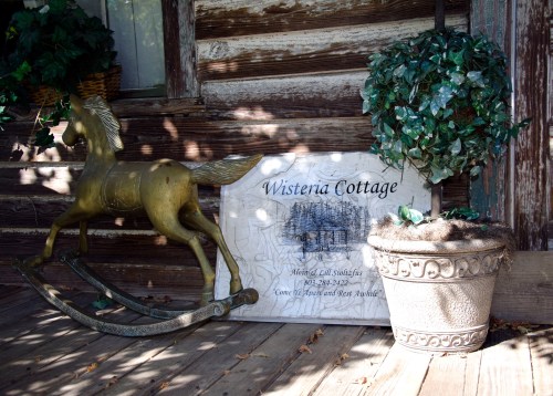A rocking horse and a potted plant beside a sign reading "Wisteria Cottage" on a rustic wooden porch.