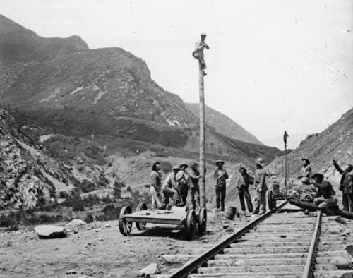 A historical black-and-white photo of workers near railway tracks, with one man climbing a pole in a mountainous landscape.