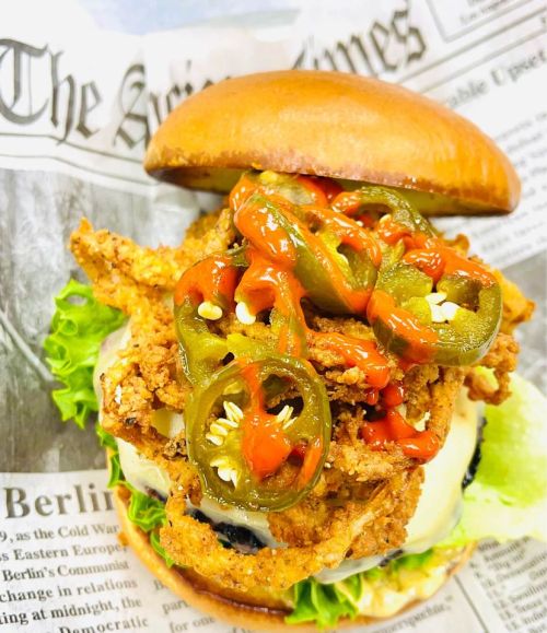 A crispy fried chicken sandwich topped with jalapeños, lettuce, and ketchup, served on a bun with newspaper wrapping.