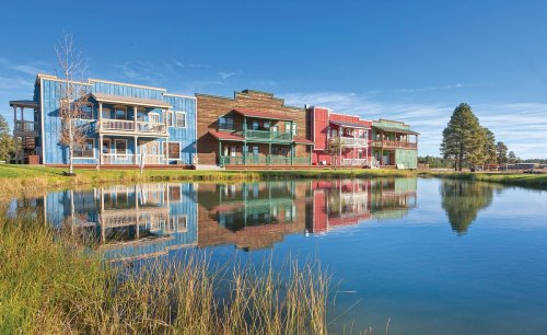 Colorful buildings reflect in a calm pond, surrounded by grass and trees under a clear blue sky.