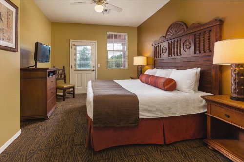 Cozy hotel room with a king bed, wooden furniture, TV, and warm-colored walls. Natural light from a window.