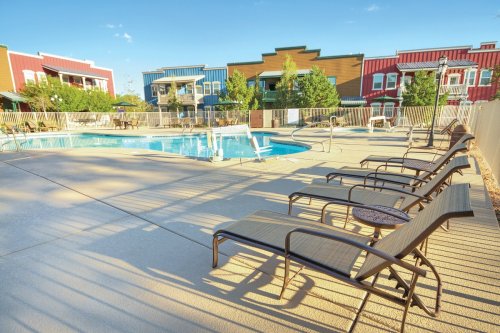 A sunny pool area with lounge chairs, surrounded by colorful buildings and trees.