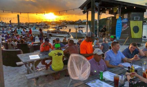 A lively outdoor restaurant by the water at sunset, with diners, boats, and a warm, inviting atmosphere.