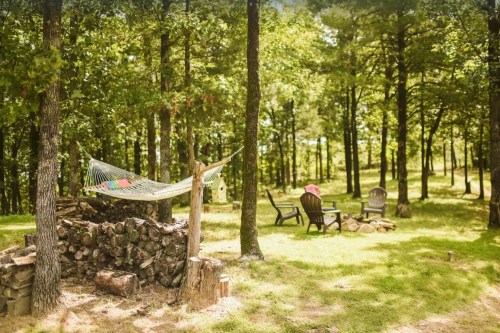 A serene outdoor scene with a hammock, fire pit, and seating area surrounded by trees and lush green grass.
