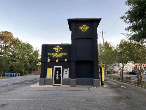 A modern coffee shop with a black exterior and yellow signage, surrounded by trees and a parking area.