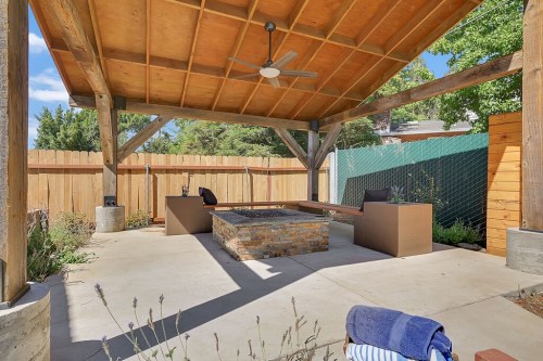 A spacious outdoor patio with a stone fire pit, wooden beams, and seating under a shaded roof, surrounded by greenery.