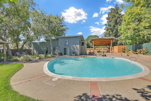 A serene backyard with a swimming pool, surrounded by greenery and a modern house under a blue sky with fluffy clouds.