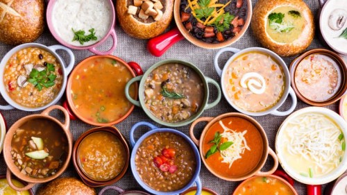 A colorful array of soups in small bowls, surrounded by bread bowls and garnishes, displayed on a textured surface.