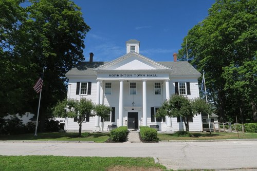 White colonial-style building with columns, labeled "Hopkinton Town Hall," surrounded by trees and a clear blue sky.