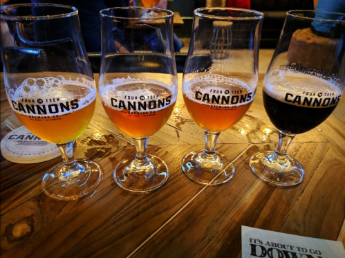 Four glasses of beer in varying colors, placed on a wooden table, with the logo "Fourteen Cannons" visible.