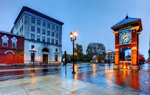 A rainy street scene featuring a clock tower and historic buildings, with wet pavement reflecting the surroundings.