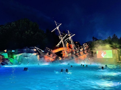 A pirate ship wreck partially submerged in a pool, with mist and a screen showing a movie in the background at night.