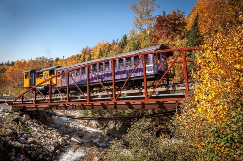 A colorful train crosses a red bridge over a rocky stream, surrounded by vibrant autumn foliage.