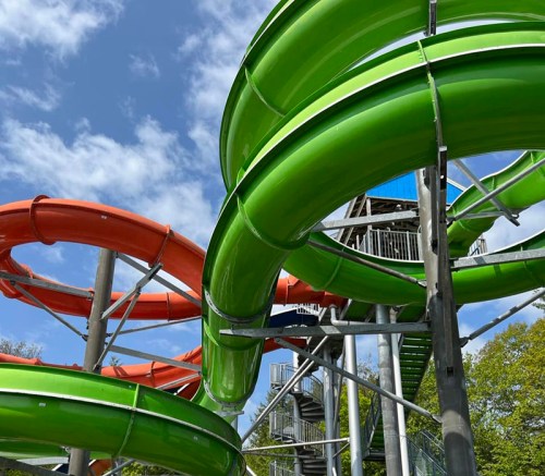 Colorful water slides in a park, featuring green and orange tubes against a blue sky with scattered clouds.